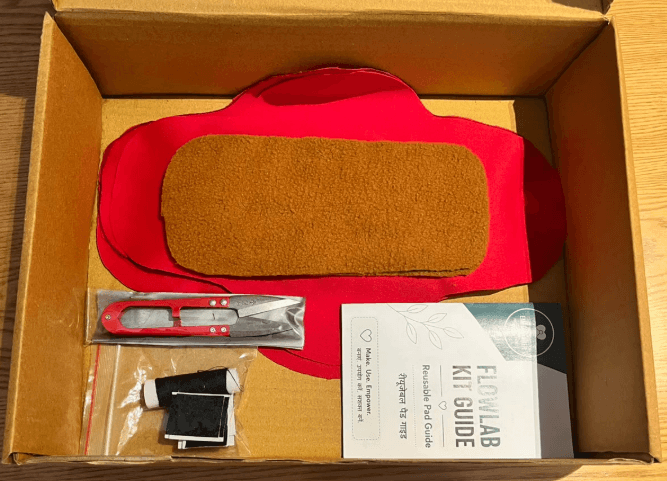 An open FlowLab Kit showing a reusable cloth pad, thread, needle, velcro, a thread cutter and the pad-making guide